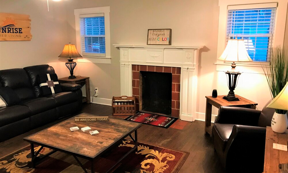 a living room area with windows, fireplace, black couch, lamps, and a wooden center table over the carpeted hardwood floor